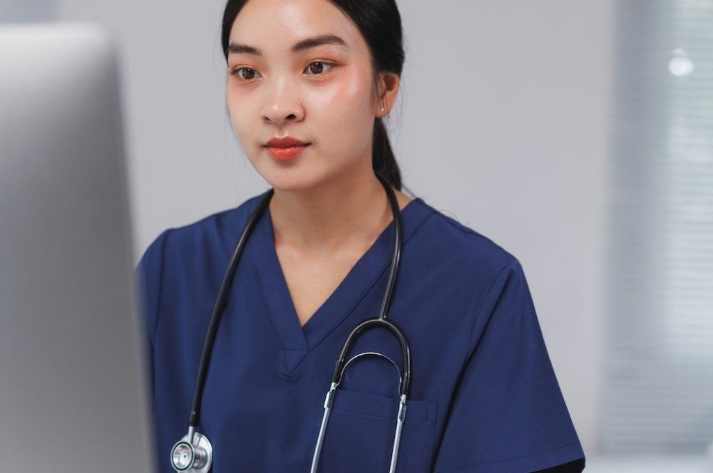 Asian female doctor wearing blue scrubs and a stethoscope, working on a desktop computer in a modern clinic setting